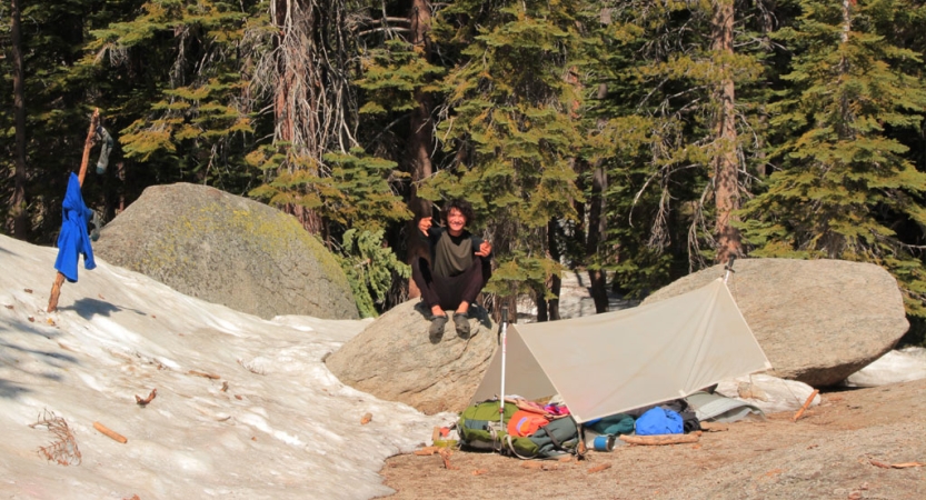 A person sits on a rock near a tarp shelter